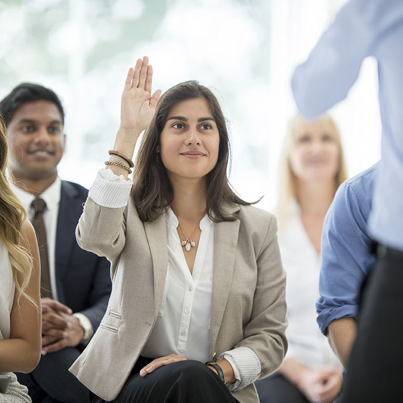 A multi-ethnic group of corporate business professionals are getting trained by the companies human resources department. They are listening to a presentation - one woman has her hand raised to ask a question.
